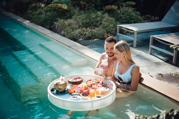 Summertime weekend in Thailand. Handsome guy with his seductive wife drinks cocktails in swimming...