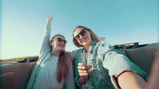 Woman with glass of champagne takes selfie with friend raising her hands up and fooling around. Young women with glasses take pictures of themselves on phone while ride convertible cabriolet