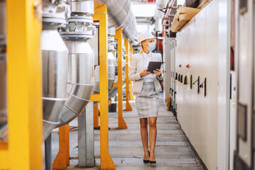 Full length of blond female boss in formal wear, with helmet on head, with protective mask walking trough heating plant and using tablet to check on machinery.