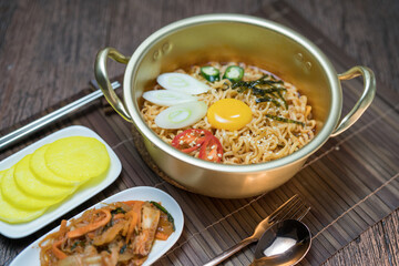 Korean style instant noodle, Ramyeon with egg yolk, chilli, kimchi vegetables pickled and radish pickles in a traditional korean noodle pot.