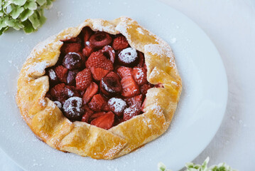 Delicious freshly baked vegan strawberry and cherry galette on white background with hydrangea flowers, close up. Sweet food, summer dessert.