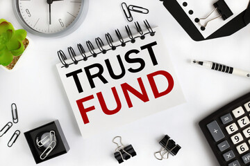 TRUST FUND. text on white notepad paper on light background near calculator, plant, table clock.