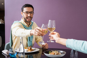 Fototapeta na wymiar Cheerful arabian man clinking wine with girlfriend near dinner in hotel room