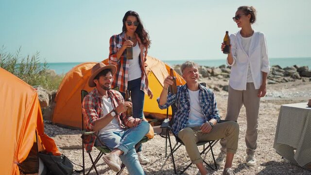 group friends travelers relaxing in camp chairs at tents in camping on sea coast. four friends on picnic drinking cold beer at hot sunny day on beach