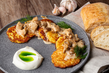 Fried potato pancakes served with mushrooms, sour cream and dill on a grey round plate. Close-up isolated on wooden background with grey linen napkin, sliced bread and garlic.