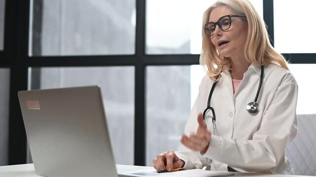 A confident female doctor, physical, therapist wearing glasses and medical gown talking with patient online, MD recommends treatment via video call, video meeting. Telemedicine