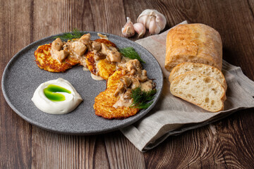 Fried potato pancakes served with mushrooms, sour cream and dill on a grey round plate. Close-up isolated on wooden background with grey linen napkin, sliced bread and garlic.