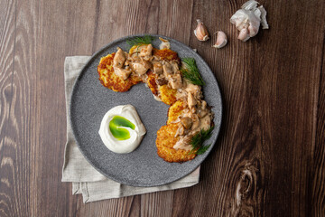 Fried potato pancakes served with mushrooms, sour cream and dill on a grey round plate. Close-up isolated on wooden background with grey linen napkin and garlic. Top view, copy space.