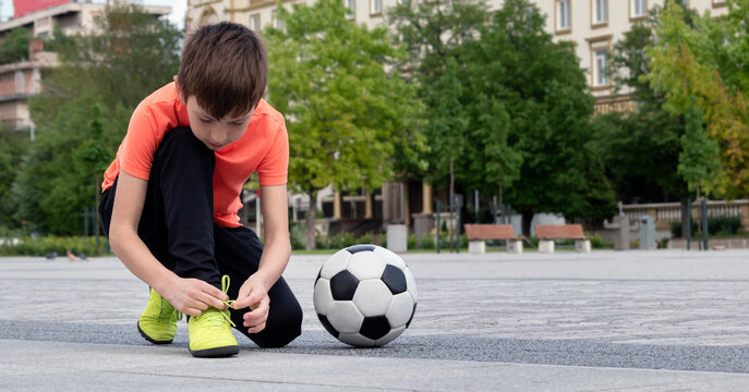 Boy soccer player tying shoelaces and soccer ball near him in town square, blurred background of cityscape in background. Sports games and leisure outside for children concept. Selective focus
