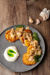 Fried potato pancakes served with mushrooms, sour cream and dill on a grey round plate. Isolated on wooden background with cloves of garlic on the table. Close-up, copy space.