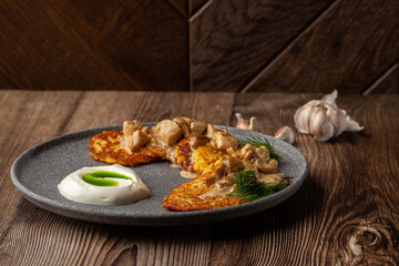 Fried potato pancakes served with mushrooms, sour cream and dill on a grey round plate. Close-up isolated dish on wooden background with cloves of garlic on the table.