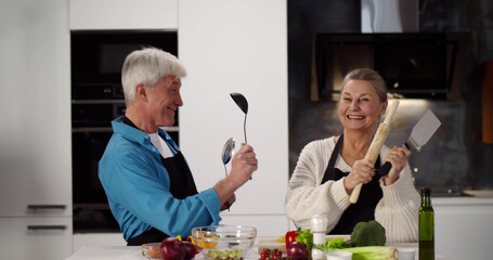 Senior happy couple in apron having fun preparing healthy food in modern kitchen at home.