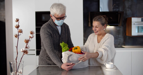 Senior man in safety mask coming home with groceries and unpacking with wife