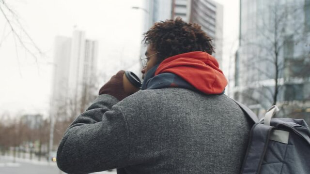 Tracking shot of young Afro-American man in outerwear drinking to go coffee and walking in city on winter day