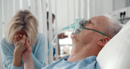 Crying woman holding hand of aged husband patient while sitting by bed at hospital