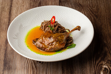 Roasted duck thigh with brown spaghetti. Served with yellow and green sauces and decorated with mint leaves and slice of chilli pepper. Close-up in a white bowl on wooden background. Isolated dish.