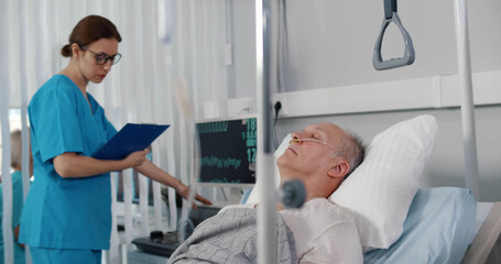 Obraz premium Doctor checking data on medical monitor while senior aged patient sleeping in bed at hospital ward