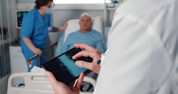 Nurse taking care of aged patient in bed while doctor examining lungs xray on digital tablet