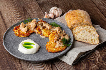 Fried potato pancakes served with mushrooms, sour cream and dill on a grey round plate. Close-up isolated on wooden background with grey linen napkin, sliced bread and garlic.