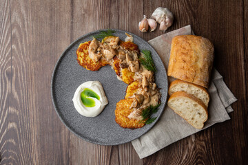 Fried potato pancakes served with mushrooms, sour cream and dill on a grey round plate. Close-up isolated on wooden background with grey linen napkin, sliced bread and garlic. Top view, copy space.