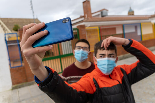 Stock photo of happy young boys wearing face mask due to covid19 taking selfie with mobile phone in the street.