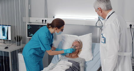 Doctor and nurse examining aged female patient in bed and putting on her oxygen mask