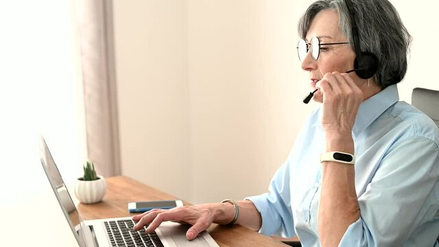 Grey-headed mature senior female office worker sitting at the desk with a laptop, wearing glasses and headset, communicating with customers and makes a marks in computer app
