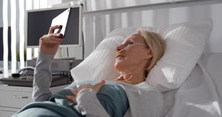 Aged female patient with breathing tube having video call on tablet pc lying in hospital bed