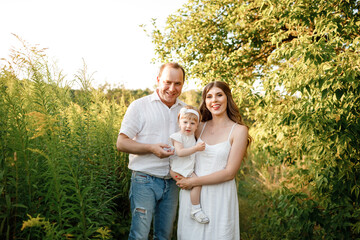 Fototapeta premium Family enjoying life together in the summer park. Happy young family outdoors. Mother, father are hugging their daughter outdoors and having fun in nature. The concept of family summer holiday.