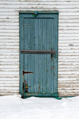 A vintage teal blue colour wooden shutter door on a white building with a mound of fresh white snow on the ground. The door has been sealed shut with a wooden plank. There's a rodent hole in the door.