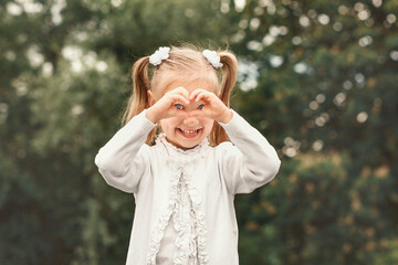 Little blonde girl in white on a green background. The child shows the heart with his hands and looks through it