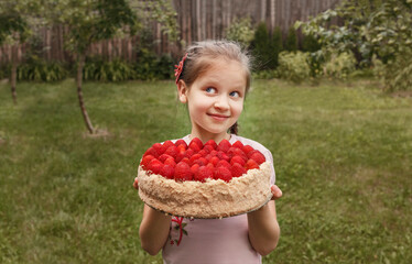 Dreamy girl holding a cake decorated with raspberries on a garden background. The child celebrates a birthday holding a holiday cake