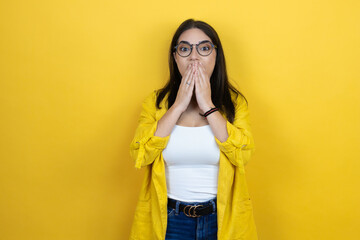 Young brunette businesswoman wearing yellow blazer over yellow background shocked covering mouth with hands for mistake. secret concept.