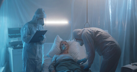 Medical workers in personal protective suits checking health of sick and patient in isolation ward