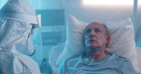 Close up of nurse wearing protective overall and face shield feeding sick aged man in hospital