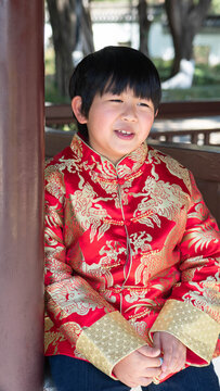 An Asian happy kid with the lovely smiling expression.A cute Chinese boy in the tradition clothes during Lunar New year festival. A child is sitting on the wooden chair at the park.