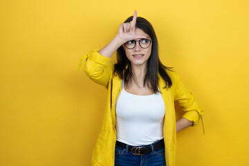 Young brunette businesswoman wearing yellow blazer over yellow background making fun of people with fingers on forehead doing loser gesture mocking and insulting.