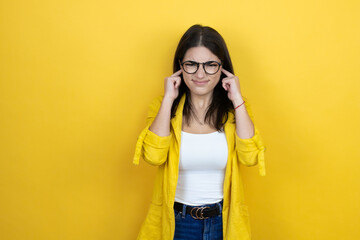 Young brunette businesswoman wearing yellow blazer over yellow background covering ears with fingers with annoyed expression for the noise of loud music. Deaf concept.