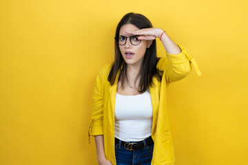 Naklejka premium Young brunette businesswoman wearing yellow blazer over yellow background very happy and smiling looking far away with hand over head. searching concept.