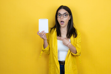 Young brunette businesswoman wearing yellow blazer over yellow background smiling, surprised and pointing blank notebook in her hand