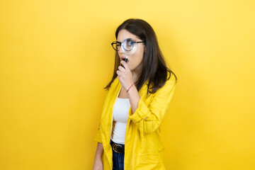 Young brunette businesswoman wearing yellow blazer over yellow background surprised looking through...