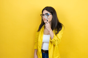 Young brunette businesswoman wearing yellow blazer over yellow background surprised looking through a magnifying glass