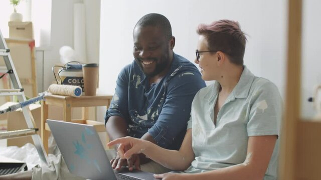 Cheerful multiethnic couple in clothes with paint stains sitting together on floor in room and discussing something on laptop screen while renovating home