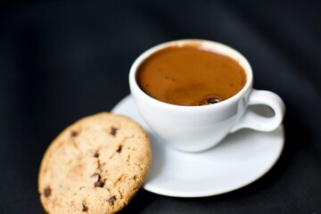 Turkish coffee in a white porcelain glass and chocolate chip cookies. Blurry and black background.