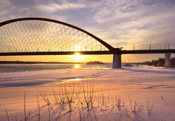 Winter morning on the Ob. Bright sunrise under the arch of the Bugrinsky automobile bridge on the frozen river bank in Novosibirsk