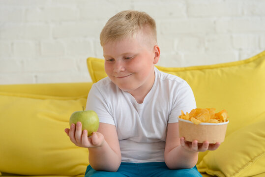 Happy fat boy looking at an apple and holding a cup of chips