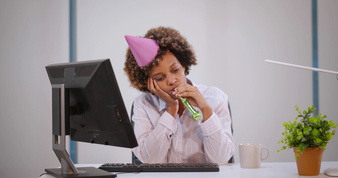 Upset woman in birthday party hat with playing pipe celebrating alone at workplace