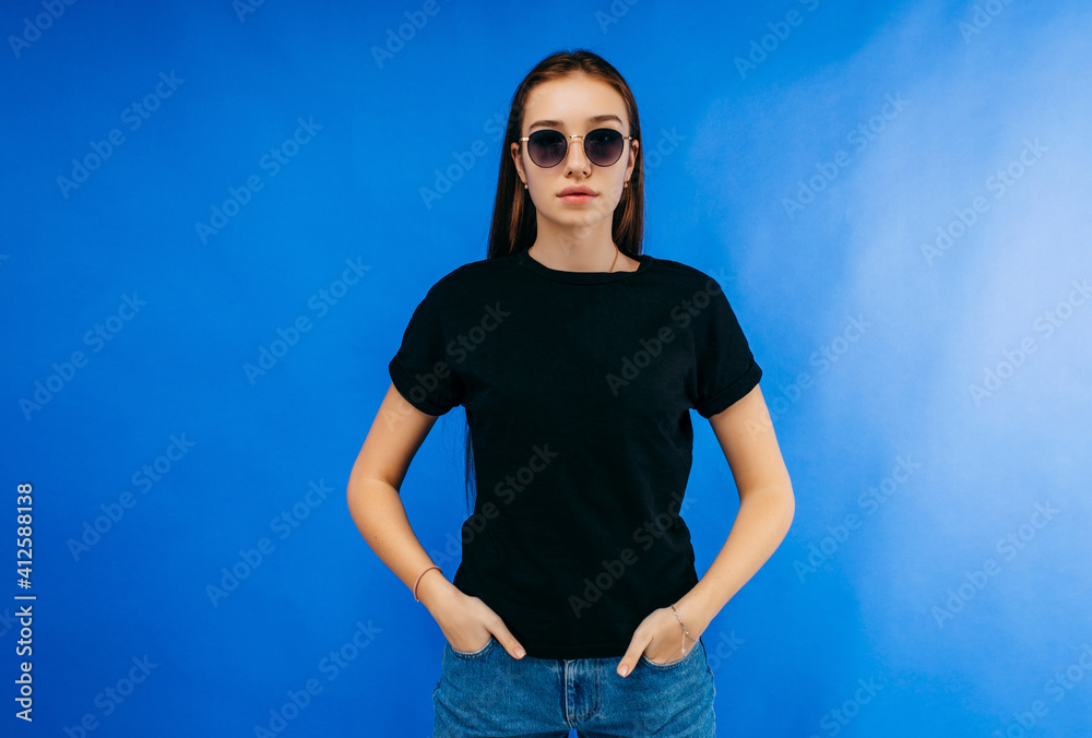 Wall mural Stylish girl in glasses wearing black t-shirt posing in studio on blue background - Wall murals
