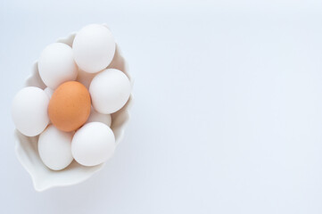 Many white eggs and one brown egg in a white dish on a light background. Copy space, top view