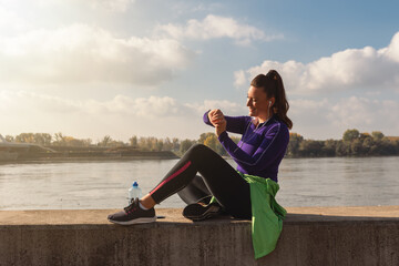 Young active beautiful handsome sporty fitness woman checking her score time on her smartwatch after outdoor training workout and taking a break from jogging and running by the river with sunrise